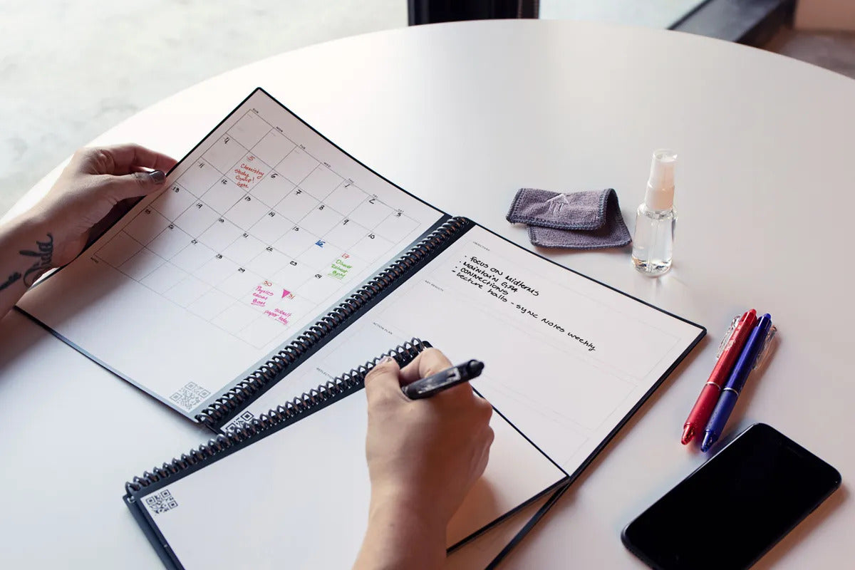 person writing in Rocketbook with Rocketbook accessories scattered on table